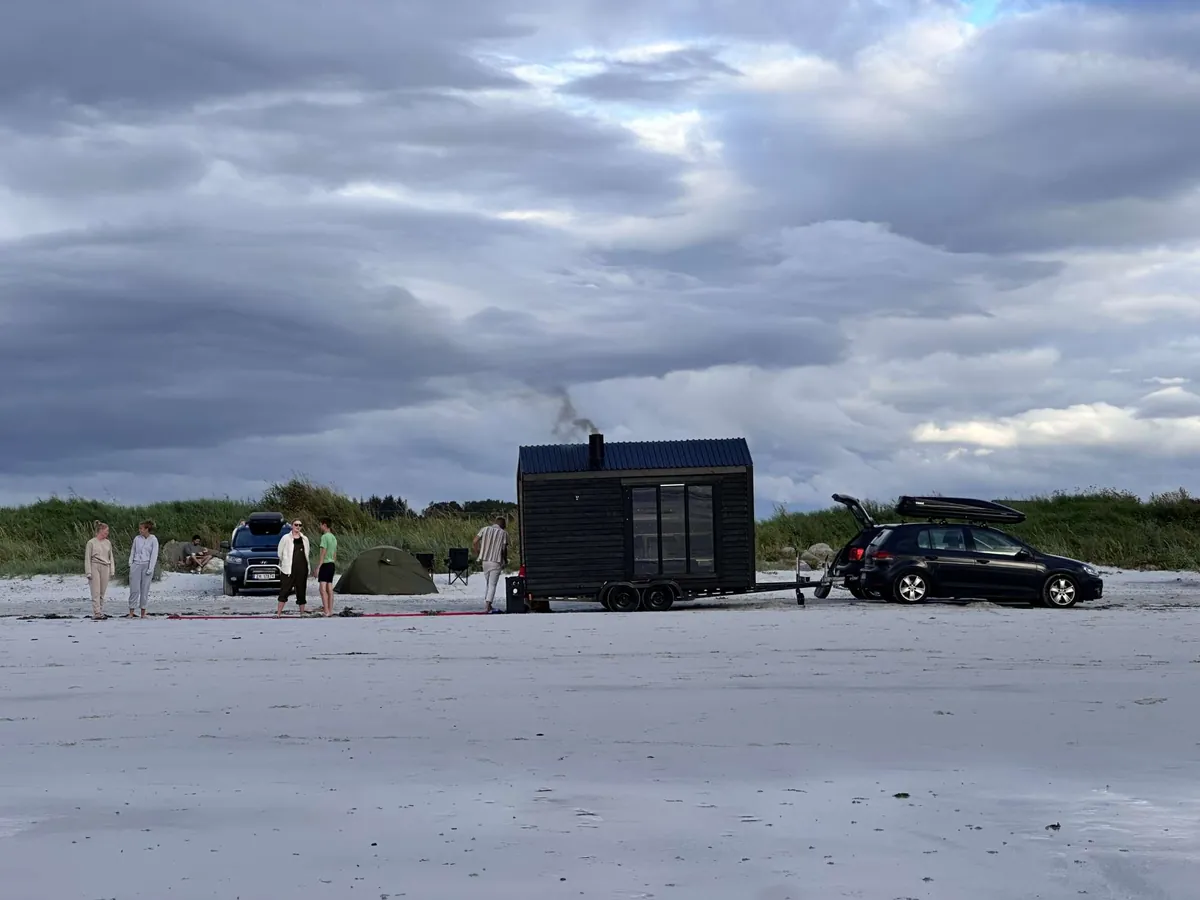 Steike – eksterior Steikes sorte badstuetilhenger parkert på en strand med gjester samlet rundt
