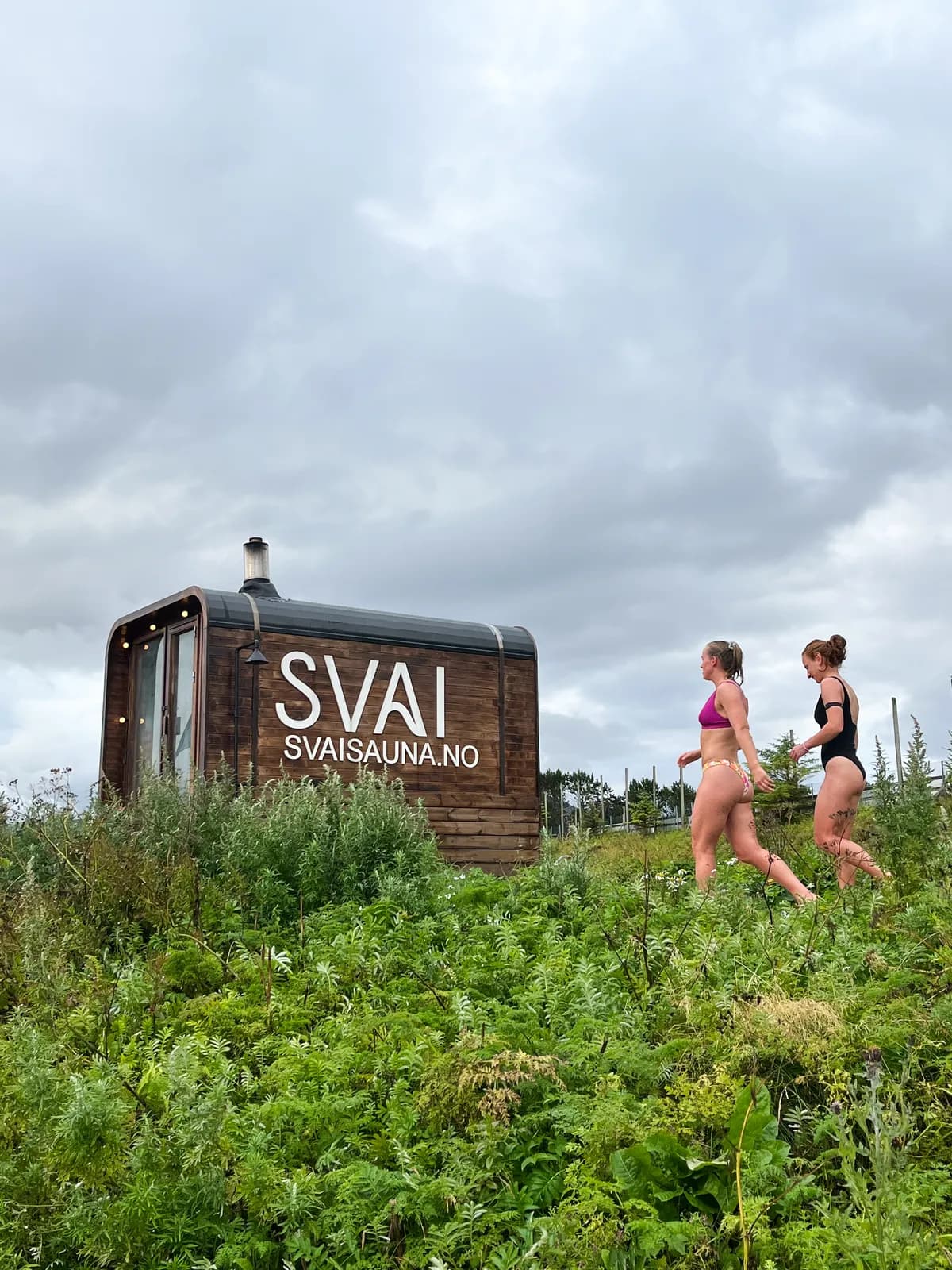 Svai Sauna Kubben, vedfyrt mobil badstue med SVAI-logo, omgitt av frodig gress med to badegjester på vei inn