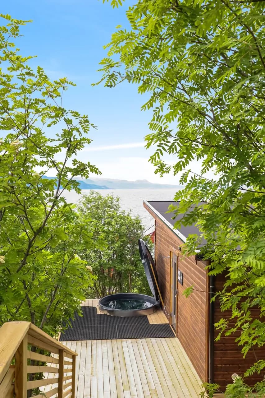 Alta Sauna – eksterior Badstuehytte i tre med kaldtbadstamp på terrasse, omgitt av trær med fjorden i bakgrunnen