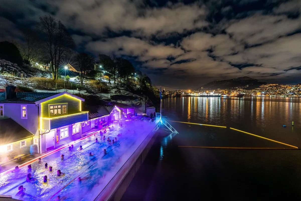 Nordnes Sjøbad i Bergen om kvelden med badende i det oppvarmede utendørsbassenget, dampsky og blålilla belysning mot Bergens bylandskap