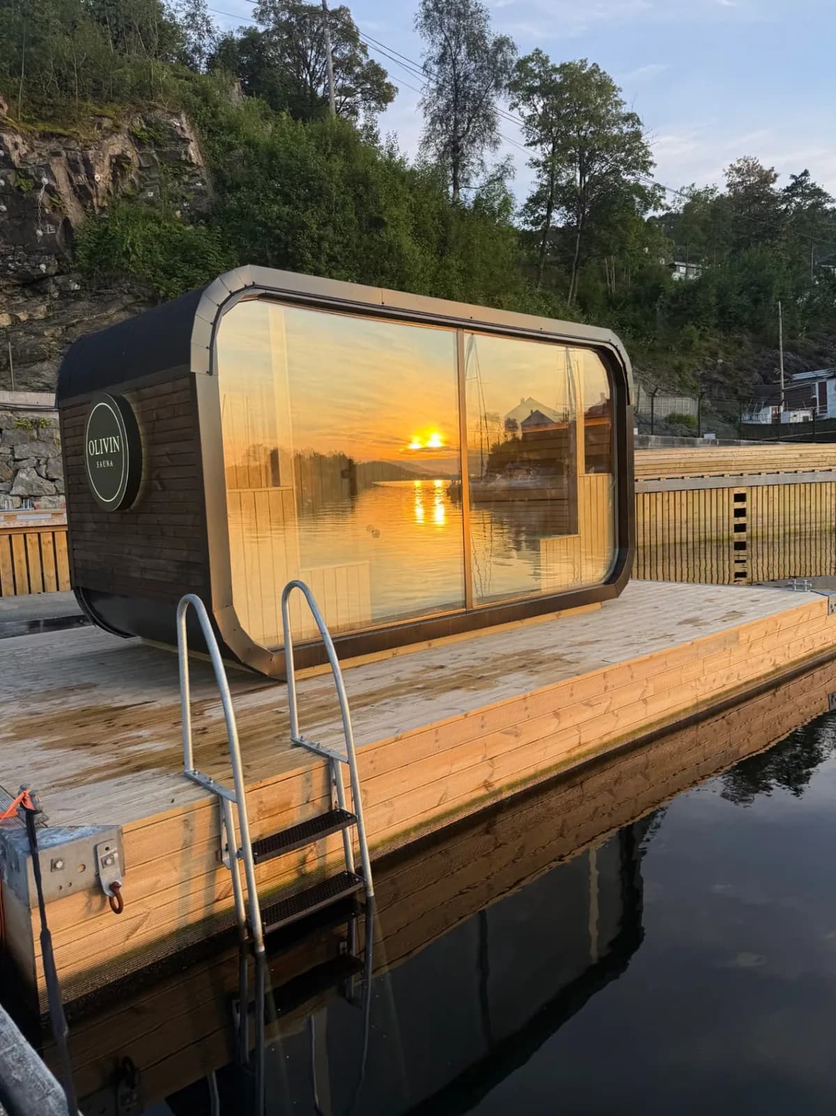 Olivin Badstue – kveld Flytende badstue på brygge ved solnedgang, med speiling av solen i store vindusflater