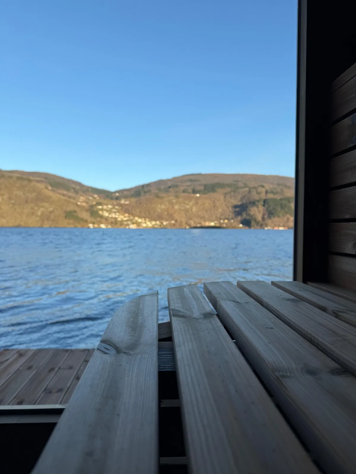 Innsiden av badstuen sett mot fjorden og fjellene gjennom det store vinduet, med ovn og bøtte i forgrunnen
