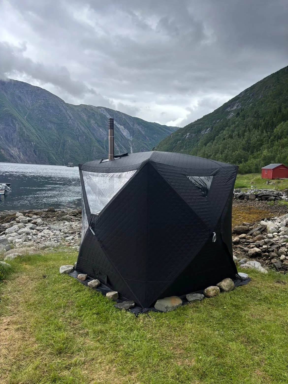 Badstutelt satt opp på gresset ved en fjord, med pipe og store vinduer