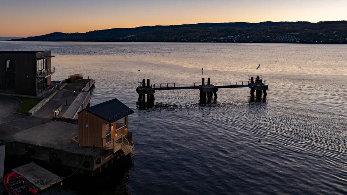 Badstuebygget på betongbrygga ved Lehmannsbrygga sett ovenfra i solnedgang, med fjorden og brygge med lyktestolper i bakgrunnen