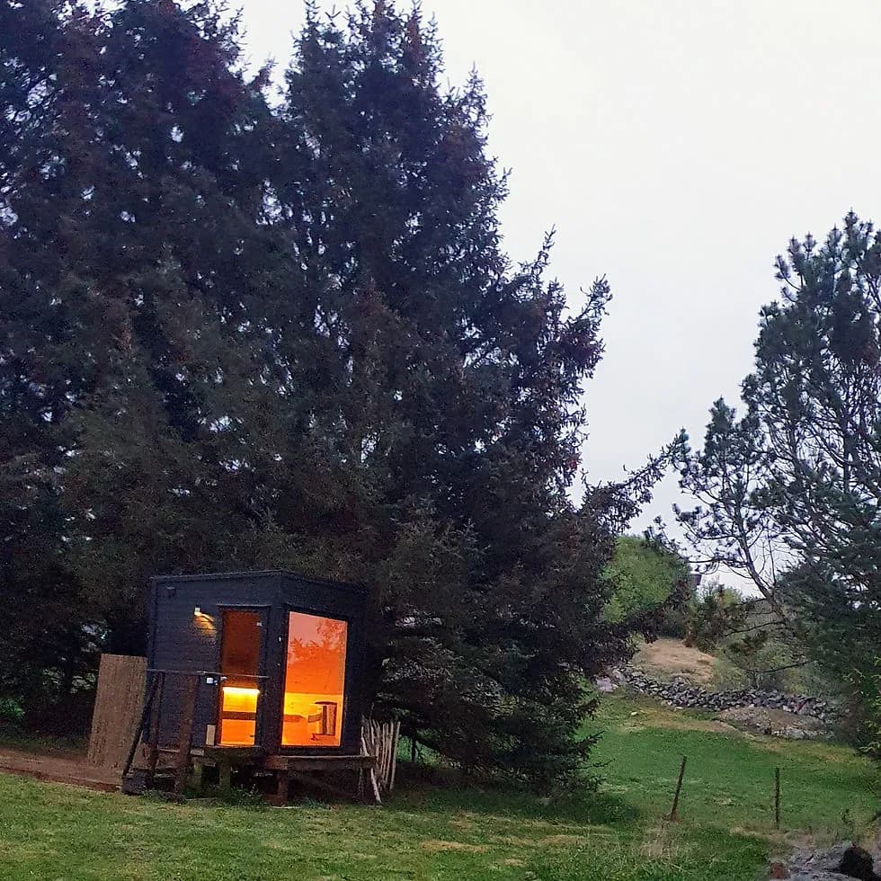 Skadberg Sauna and Glamping – eksterior Badstuehytten på Skadbergsanden i skumring, opplyst innvendig, omgitt av høye grantrær