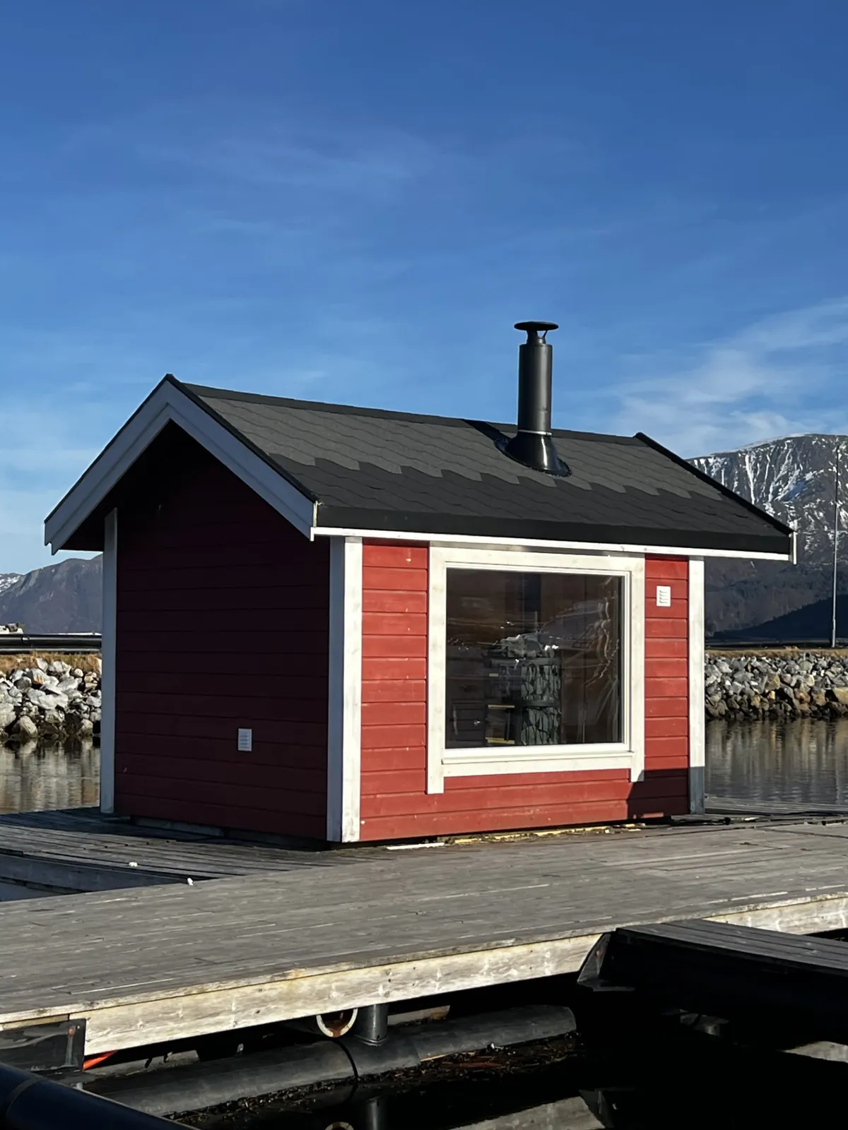 Den flytende badstuens rødmalte eksteriør med sort tak og pipe, liggende på en brygge med fjell i bakgrunnen
