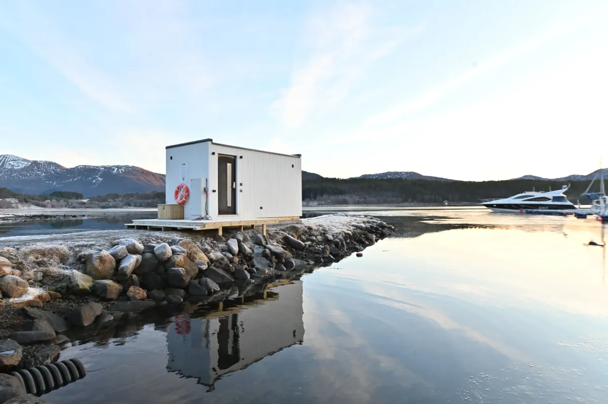 OPPKOK Sauna Elnesvågen – eksterior Hvit badstuecontainer plassert på steinet molo ved sjøen, med fjell og lystbåter i bakgrunnen ved soloppgang/solnedgang