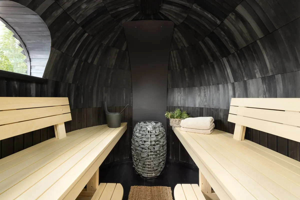 Mjøsli Sauna Experience – interior Interiøret i badstuen med en elektrisk HUUM-ovn, bøtte, håndklær og trebenker under et hvelvet, sotsvart tak