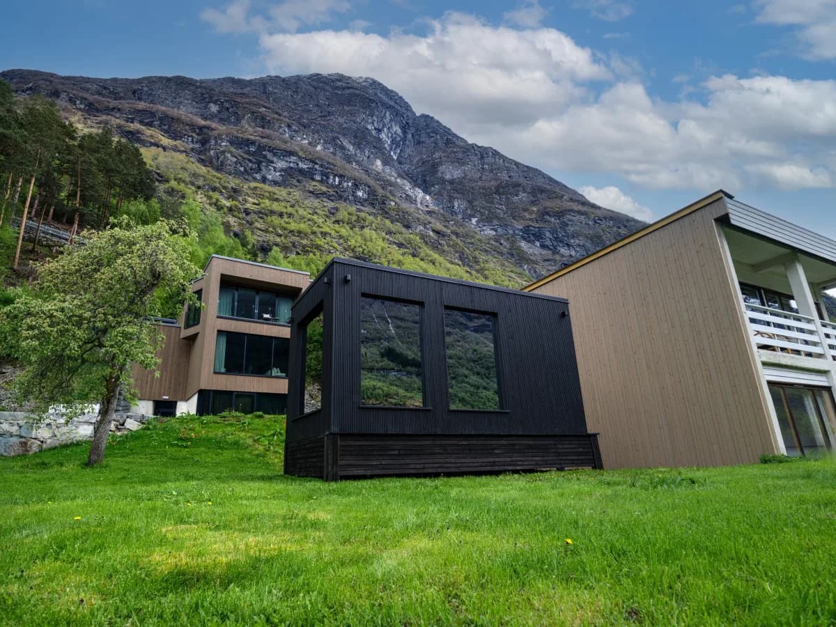 ROA Badstue Geiranger sett utenfra med fjell og Grande Fjordhotell i bakgrunnen