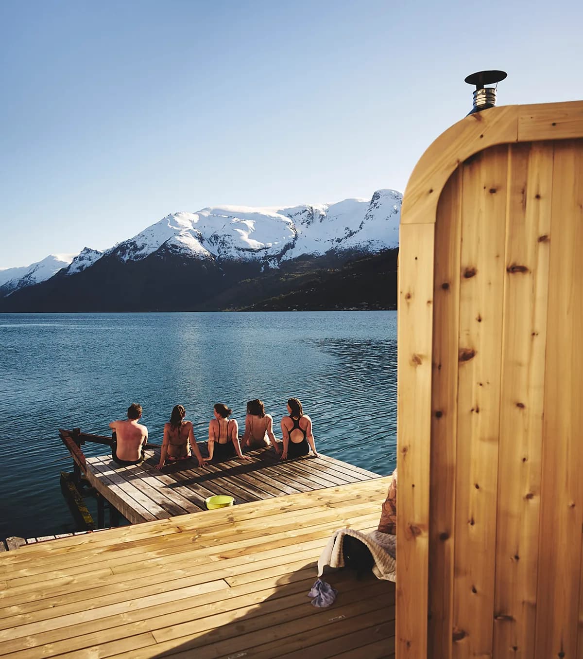 Heit Sørfjorden Sauna i Hovland med gjester på brygga og fjordutsikt mot snødekte fjell