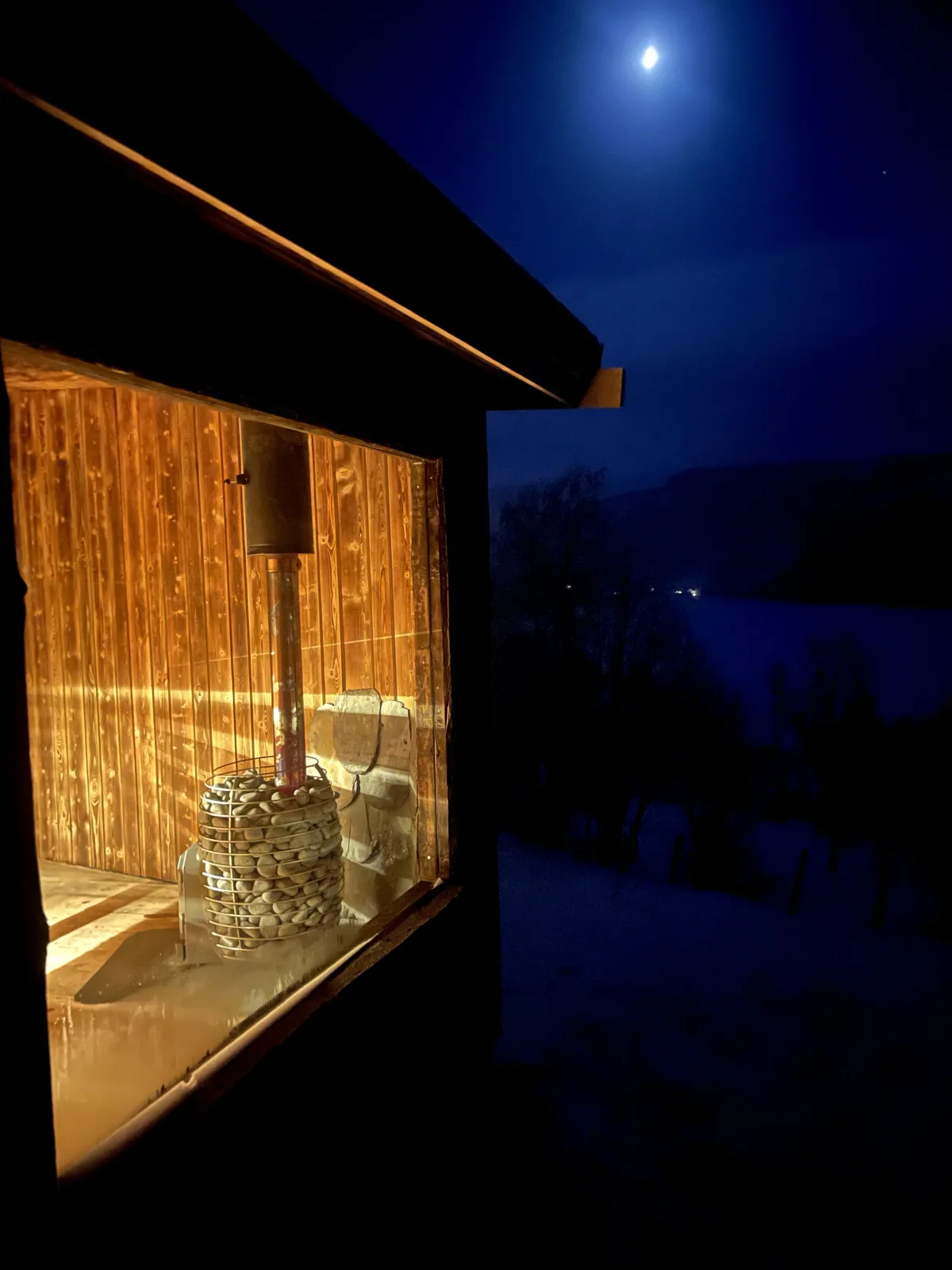 Badstogo badstue i Lomen sett utenfra om kvelden med opplyst saunaovn og måneskinn over vinterlandskap