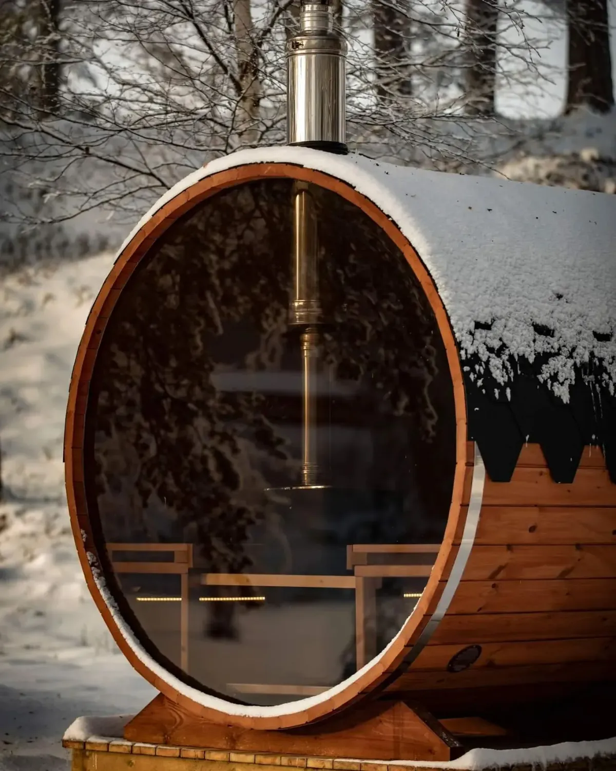 Heksas Kjøkken Sauna tønnebadstu med stort glassvindu og pipe i snødekt vinterlandskap ved Hogga sluse i Lunde