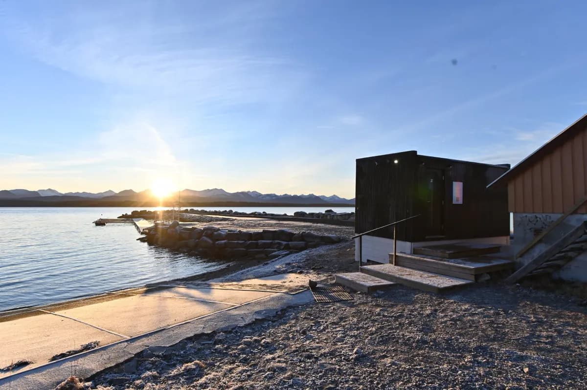 OPPKOK Sauna Lubbenes – eksterior Badstuens sorte eksteriør på strandkanten ved solnedgang, med brygge og fjell i bakgrunnen