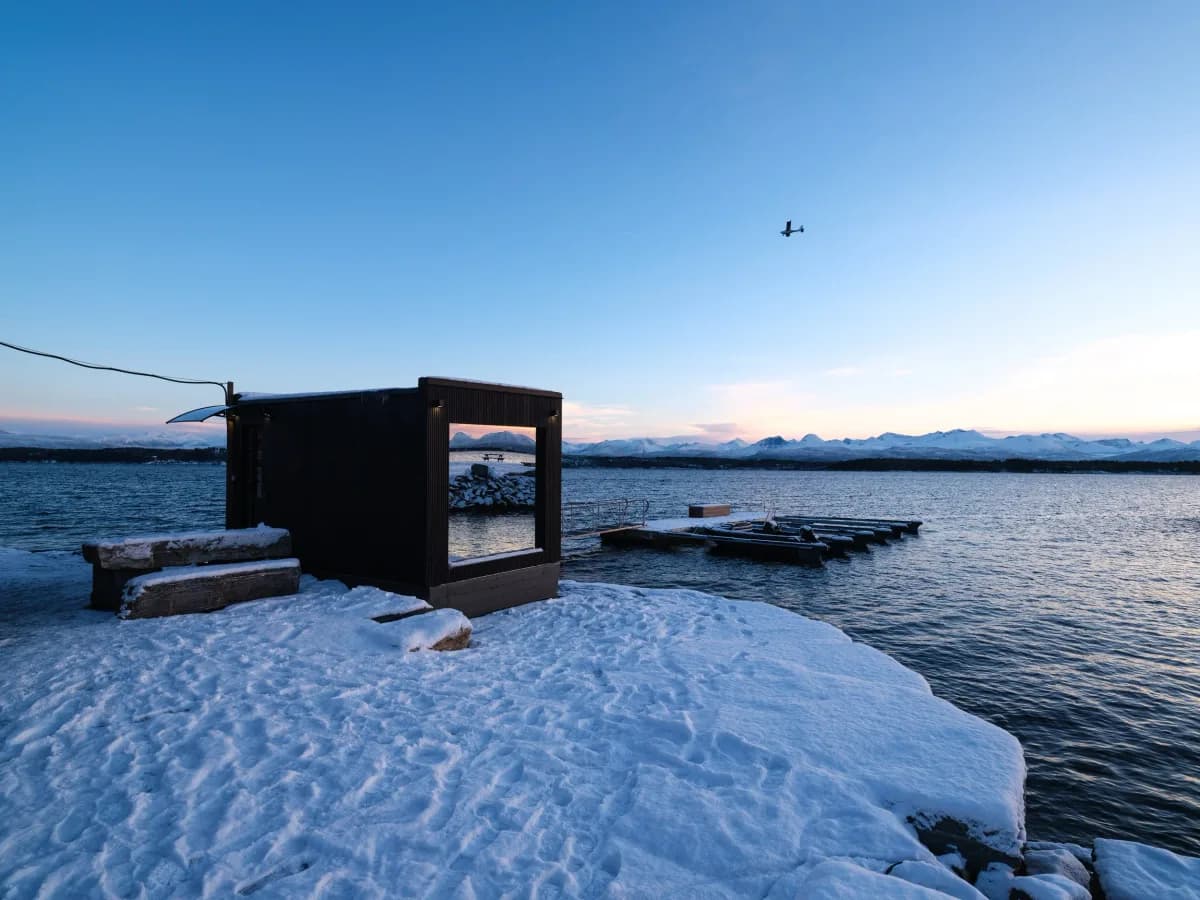ROA Kviltorp badstue i Molde, svart kubisk bygning ved fjorden med snødekt landskap og fjell