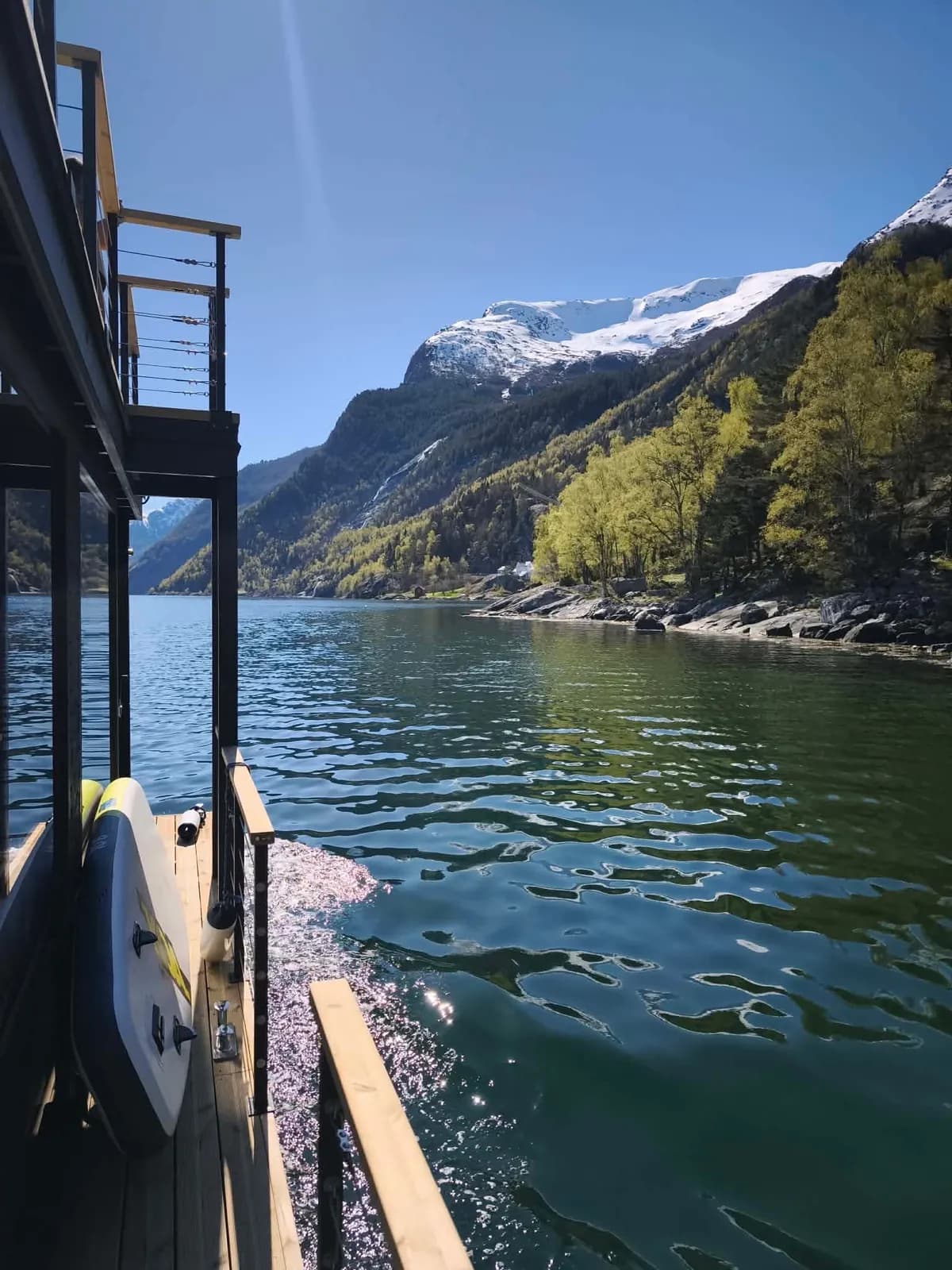 H2Odda – utsikt Utsikt frå den flytande badstua mot fjorden og snødekte fjell, med SUP-brett på dekk