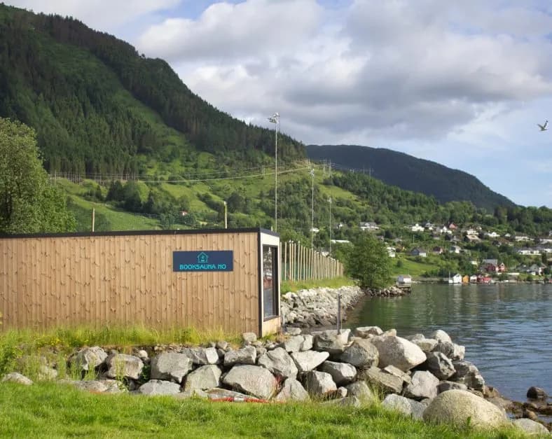 Badstuebygget i tre ved sjøen i Rosendal sentrum, med skilt 'BOOKSAUNA.NO' og fjell i bakgrunnen