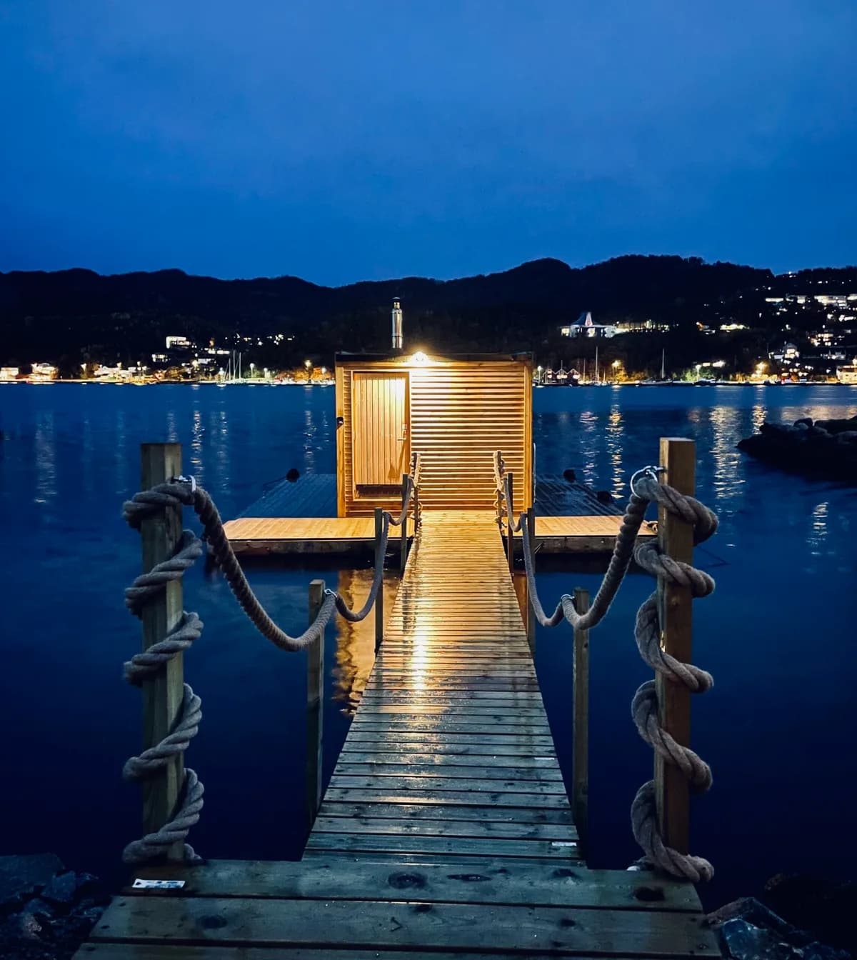 Flytende badstue BookSauna Saga i Sandnes opplyst om kvelden med gangbro over vannet