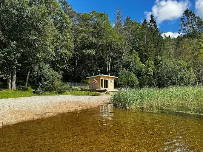 Sauna Med Sjarm badstue ved Bråsteinvatnet i Sandnes sett fra vannkanten med sandstrand og grønn natur