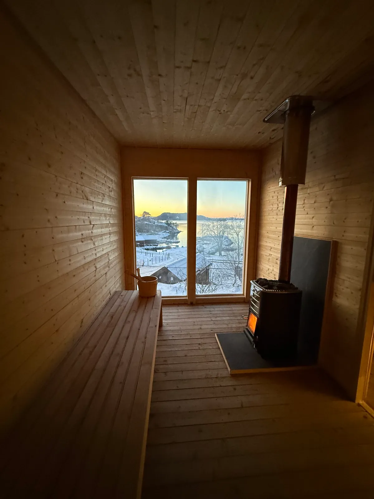 Sørvest Sauna – interior Interiør i badstuen med vedovn og utsikt gjennom vinduet mot sjøen i vintersolnedgang