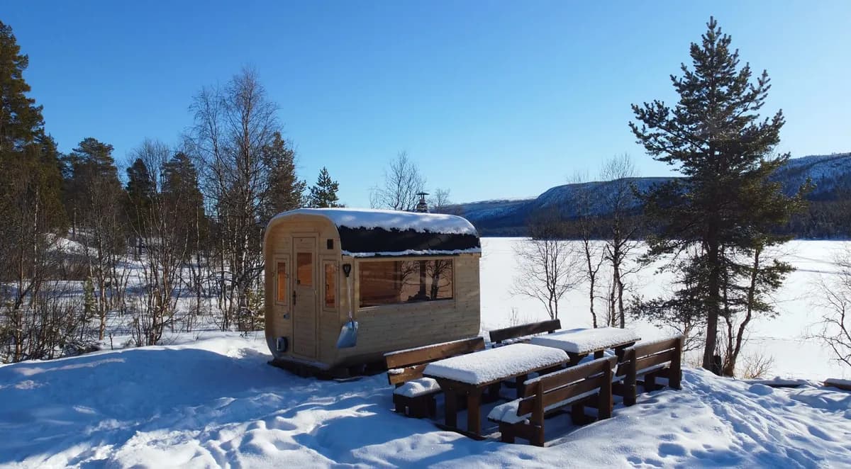Bygdebadstua i Skurdalen – tønnebadstu i lyst tre ved islagt vatn med snødekte fjell og sittebenker ute
