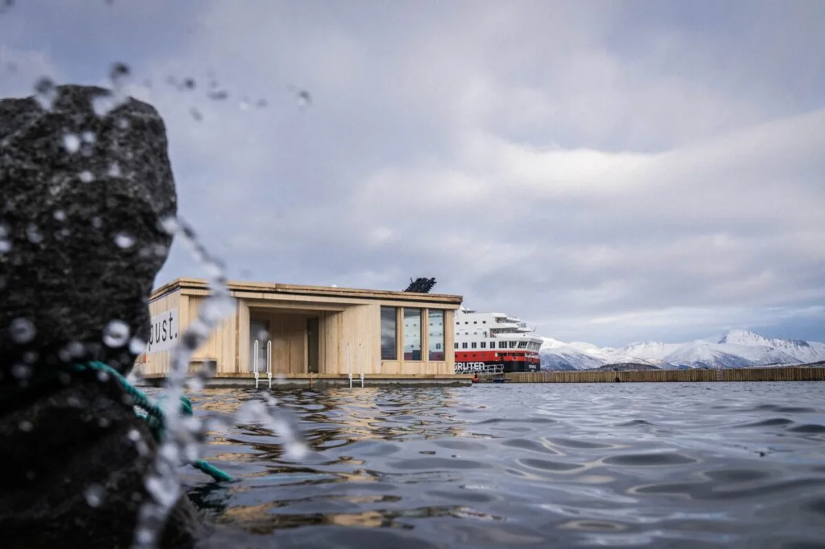 pust. Sortland badstue sett fra sjøen med Hurtigruten og snødekte fjell i bakgrunnen
