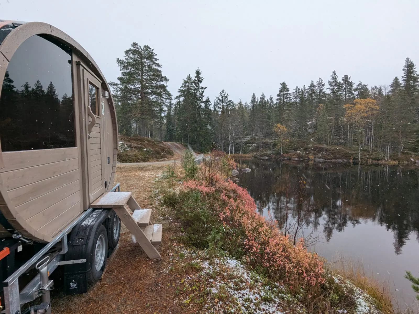 Blystad Mobile Badstu, en vedfyrt tønnebadstu på tilhenger, parkert ved et stille skogsvann en grå høstdag