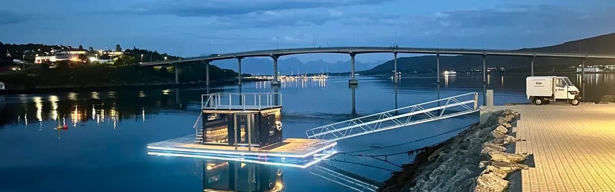 Dypp badstuflåte opplyst om kvelden i Stokmarknes havn med bro i bakgrunnen