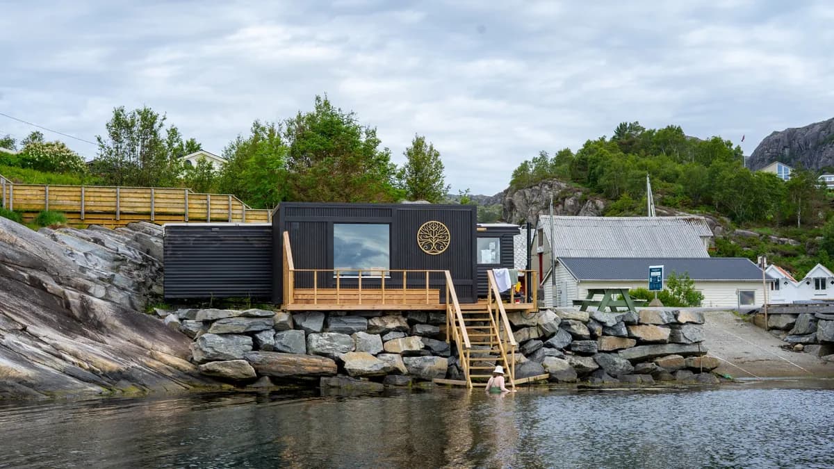 Sort badstuebygg på svaberg ved sjøen, med en person som bader ved trappen ned til vannet