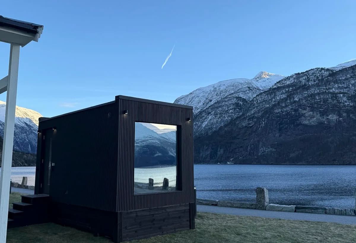 ROA Badstue Valldal sett utenfra med fjord og snødekte fjell i bakgrunnen, Sylte