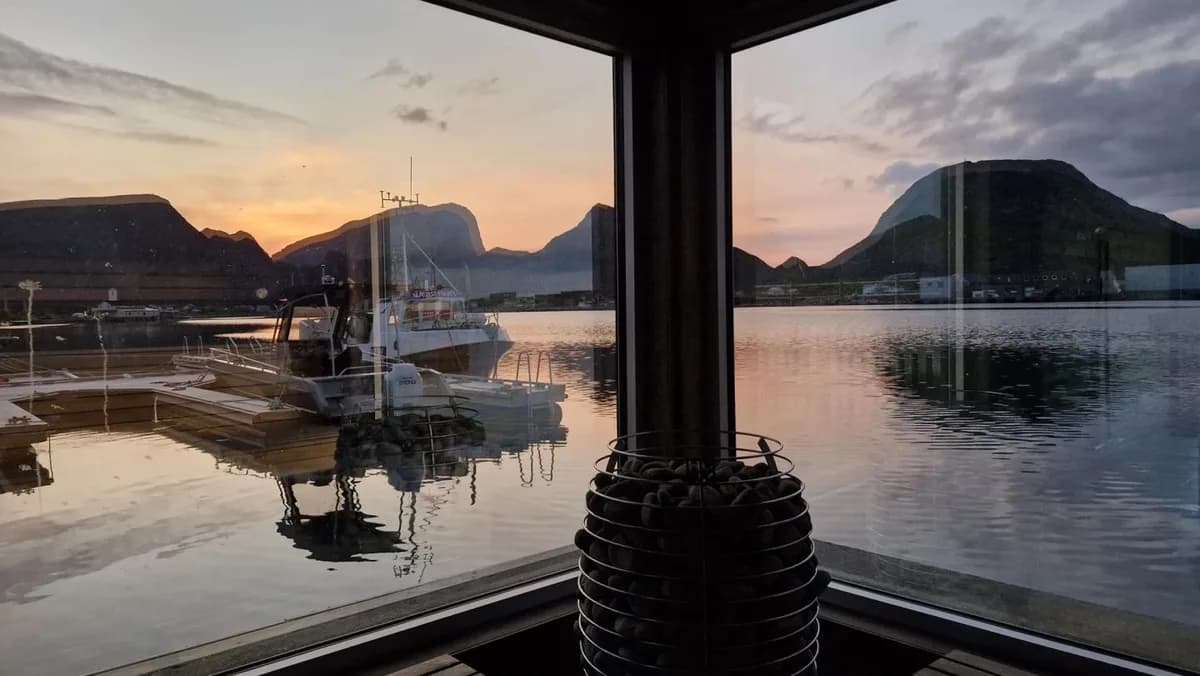 Interiør av FLYT Sauna på Værøy med saunaovn og utsikt over havn og fjell i solnedgang