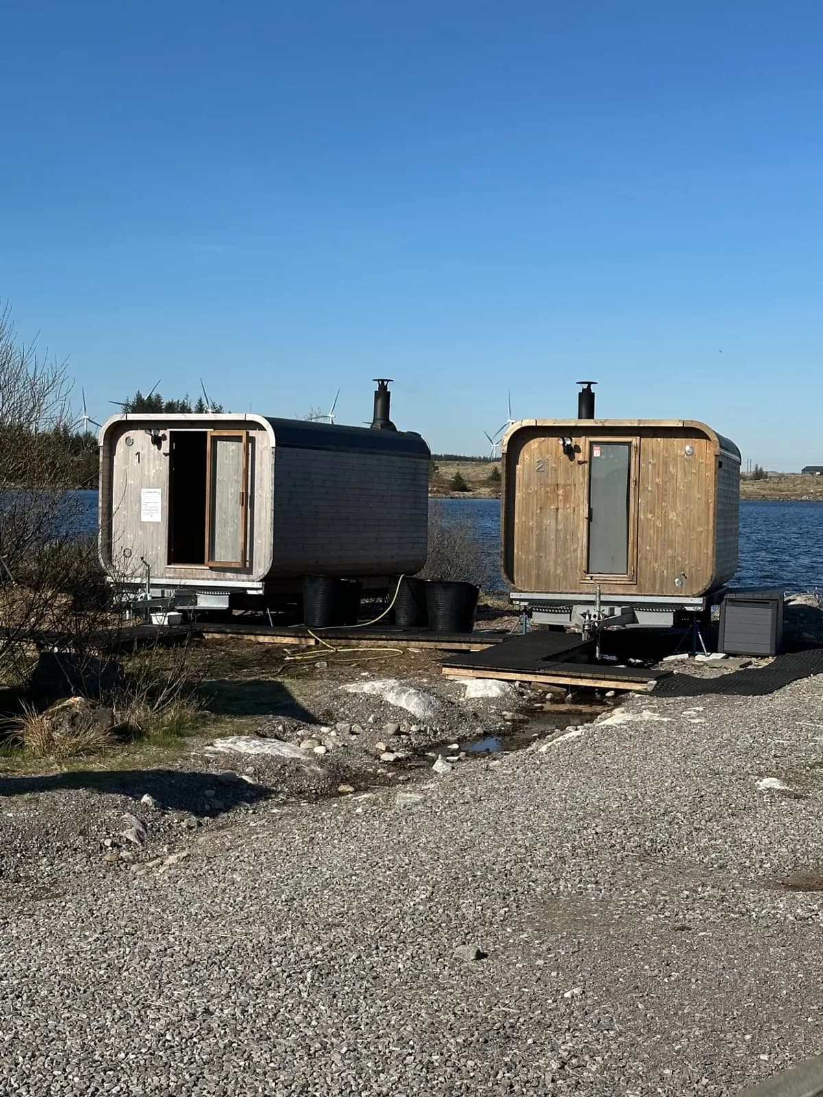 To vedfyrte badstuer på hjul ved bredden av Holmavatnet, med vindturbiner i bakgrunnen