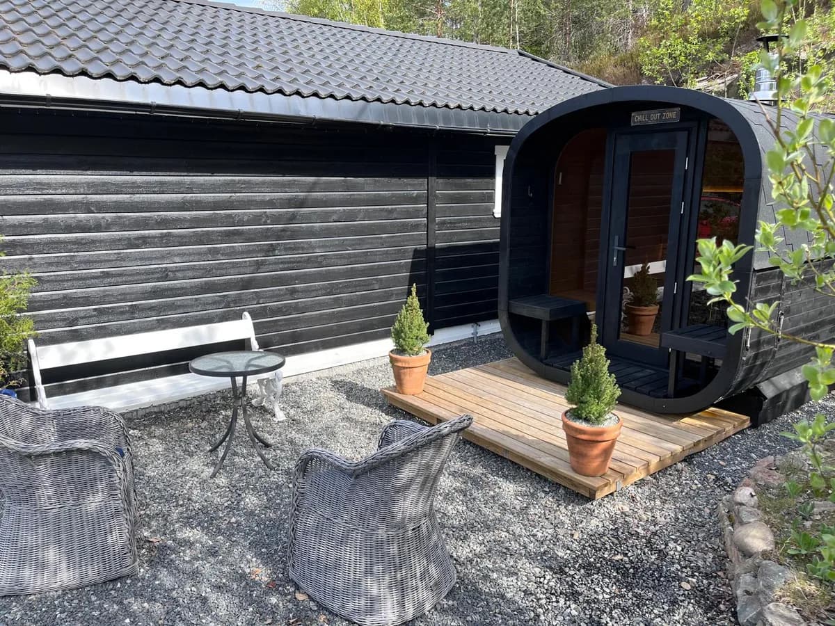 Vegårstue – eksterior Sort tønnebadstue merket «Chill Out Zone» på en treterrasse ved et hus, med hagemøbler foran